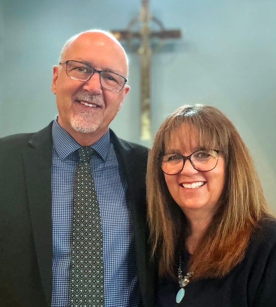 Pastor Jeff Whitt and his wife Jill, poses together in front of a cross in a church setting.
